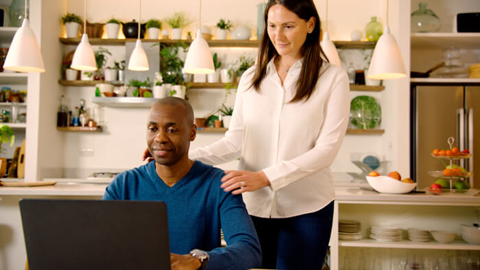 Bilde av mann som sitter p&aring; kj&oslash;kkenet med kona si st&aring;ende ved siden av ham, hvor begge ser p&aring; en b&aelig;rbar PC