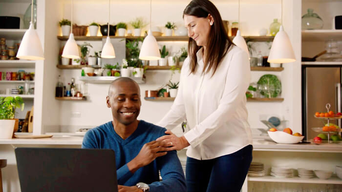 Bilde av mann som sitter og smiler, og kona hans som st&aring;r ved siden av ham p&aring; kj&oslash;kkenet og smiler ned til ham