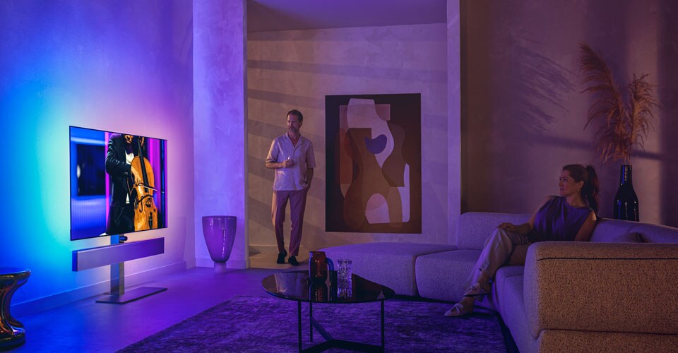 Mann og kvinne i en stilig stue som ser på en Philips Ambilight-TV som viser en celloopptreden