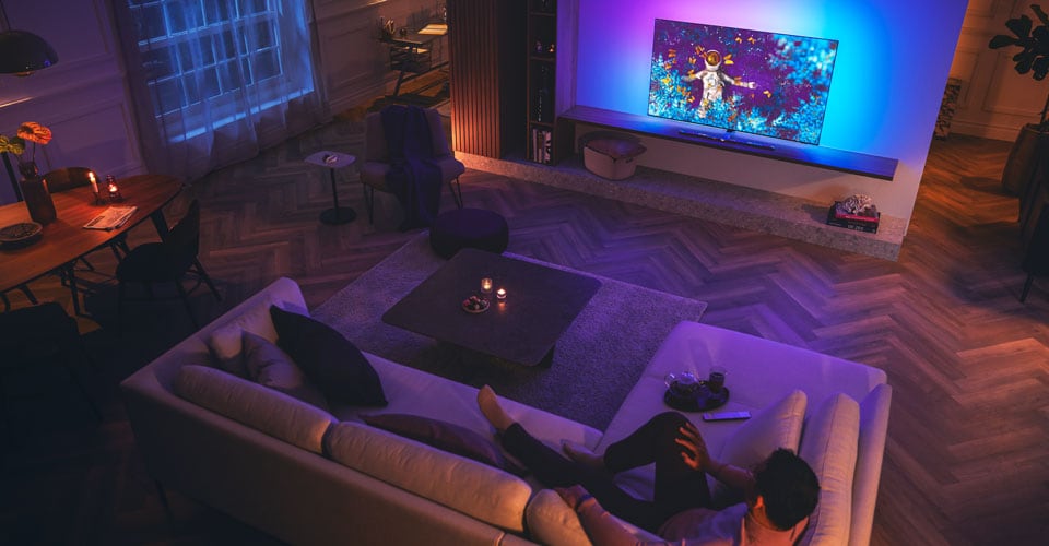 Mann slapper av på en sofa i en svakt opplyst stue og ser på en Philips Ambilight-TV