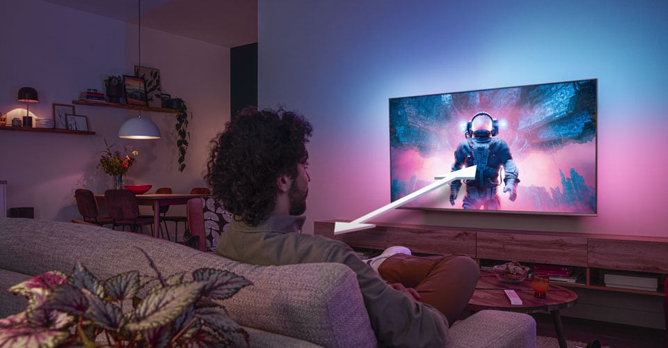 Mann sitter i en sofa og ser på en Philips Ambilight-TV som viser en fargerik astronautscene, med en pil som indikerer optimal seeavstand