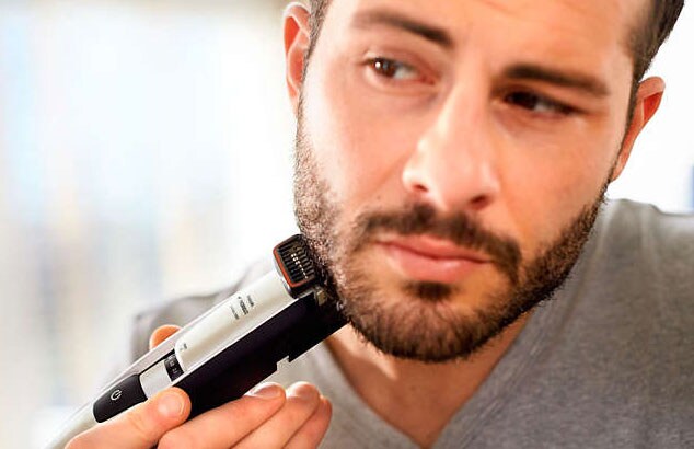 Nærbilde av en mørkhåret skjeggete mann i grå t-skjorte som bruker barbermaskin på kjevelinjen.