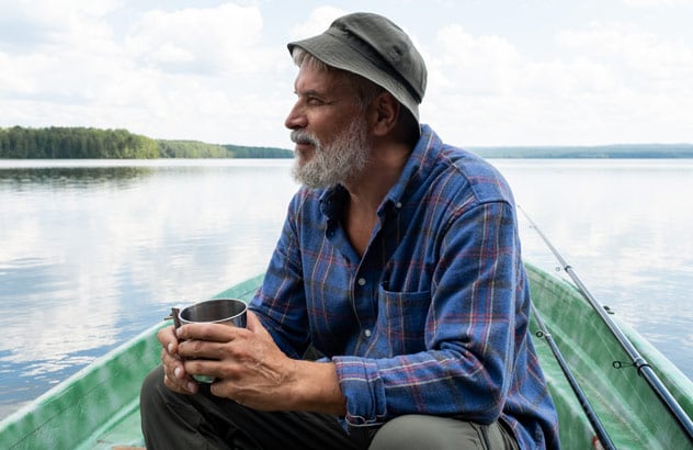En eldre mann med grått skjegg, fiskerlue og blårutet skjorte sitter på en båt og ser ut i det fjerne