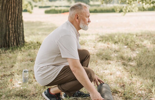 Sideprofil av en mann med grått hår og hvitt skjegg som ruller ut en treningsmatte i en eng