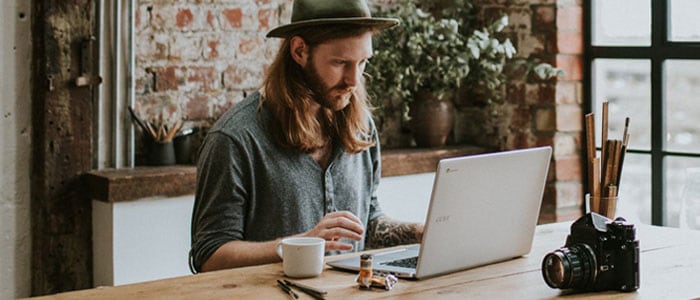 Ung mann med langt hår og helskjegg sitter på et hipt sted og ser på en laptop foran seg.