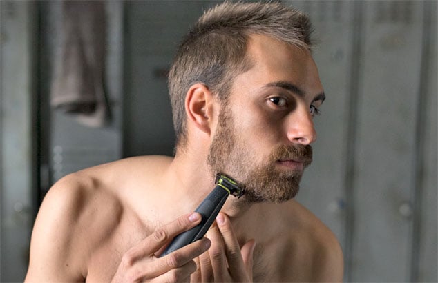 En mann trimmer det rufsete skjegget sitt ned til skjeggstubber ved hjelp av en trimmer med spesielt tilbehør.