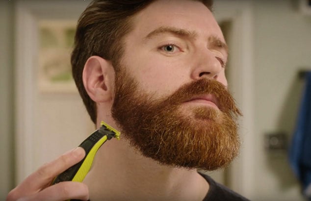 En mann trimmer det rufsete skjegget ned til skjeggstubb ved hjelp av en trimmer med et spesielt feste.