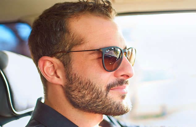 En mann kjører bil i svarte solbriller, han har et helbrunt skjegg og bart og kort hårklipp.
