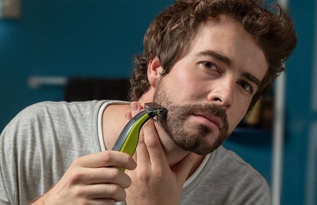 En ung mann bruker en trimmer med et spesielt tilbehør for å trimme et rufsete skjegg.
