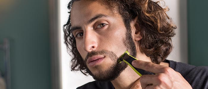 En langhåret mann bruker en trimmer for å barbere skjegget og forme barten.