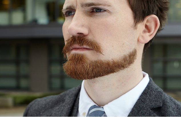 Mann med et nøyaktig barbert rødhåret chinstrap-skjegg i en mørkegrå dress ser ut i det fjerne.