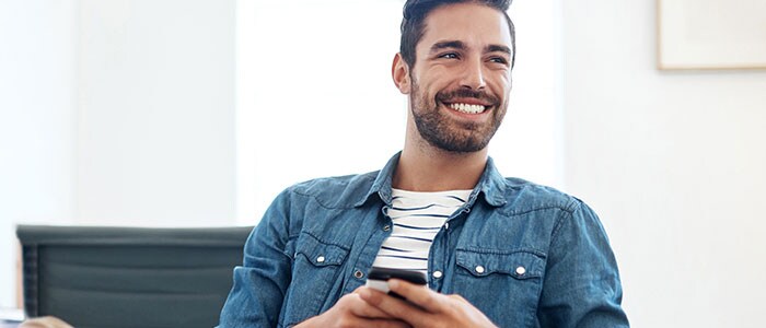 En mann med skjeggstubber og iført en denimskjorte holder en telefon mens han smiler.
