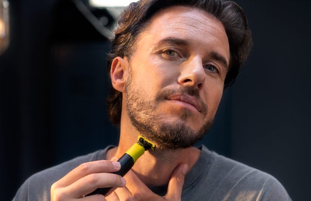 Nærbilde av en mann som trimmer kanten av det mørke skjegget sitt med Philips OneBlade foran et speil