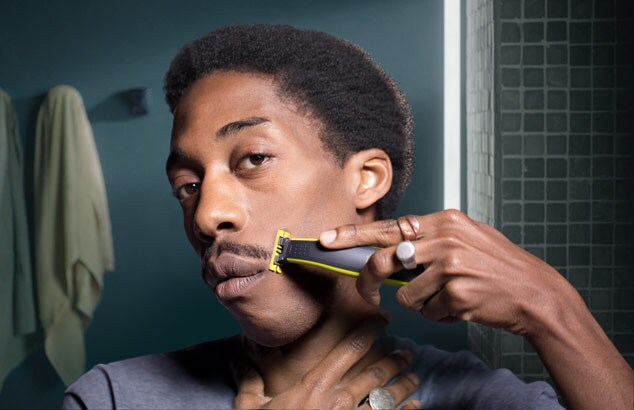 En ung svart mann trimmer barten med Philips OneBlade foran et speil på et bad.