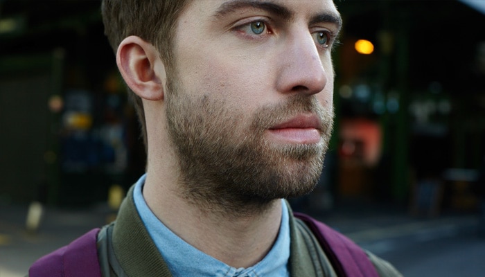 Brunhåret mann med 1 cm skjegg som dekker ansikt og hals, står i en gate iført lilla ryggsekk.